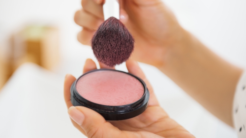 A makeup brush picks up a soft pink powder blush, ideal for fair to light skin tones